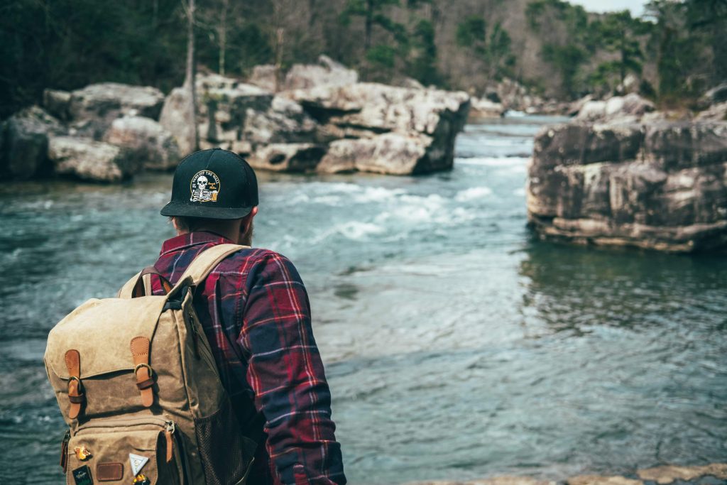 A man in plaid explores a scenic river with rocky surroundings, capturing the essence of adventure with his bagpack